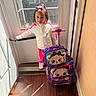 child, girl, door, window, suitcase, luggage, cat_face, pink, white_jacket, floor, sunlight, indoor, handle, car_outside, hair_bow, wood_floor, standing, casual_clothing, person, daylight