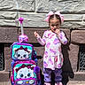 child, girl, backpack, rolling_luggage, cartoon, cat, pink, purple, bows, shoes, outdoor, stone_wall, sunlight, shadow, clothing, person, cute, accessory, toy, standing