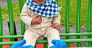 Angel is registered to the contest to win money with this photo: baby, bib, blue_hat, checkered_pattern, child, clothing, cute, footwear, grass, green_metal_structure, happy, outdoor, park, person, playground, red_floor, seated, smiling, toddler, trees