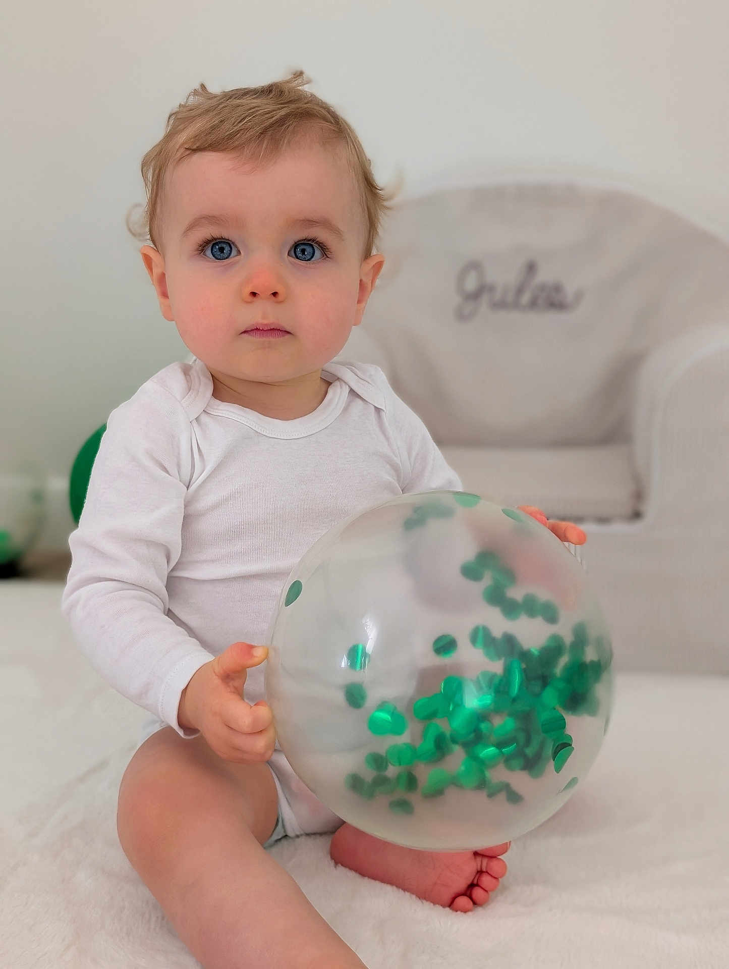 Jules a rejoint le concours — aidez-le/la à gagner de superbes lots ! baby, child, balloon, confetti, white_clothing, sitting, blue_eyes, soft_surface, indoor, curly_hair, foot, hand, chair, name_embroidery, blurred_background, cute, portrait, infant, toy, playful