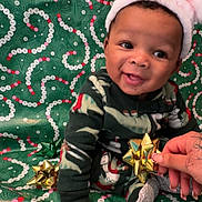 Kayden is registered to the contest to win money with this photo: baby, child, smiling, santa_hat, holiday, christmas, gift_bow, festive, pajamas, green_background, tattooed_hand, sitting, happy, cute, portrait, celebration, indoors, decor, colorful, fun