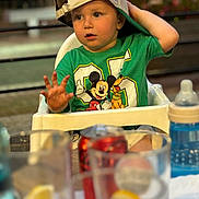 Simon a rejoint le concours — aidez-le/la à gagner de superbes lots ! toddler, child, high_chair, cap, mickey_mouse, tshirt, green_clothing, baby_bottle, table, glass, drink, blurred_foreground, outdoor, daylight, person, cute, curious, sitting, hand, face