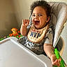 baby, bib, cheek, child, clothing, curly_hair, feeding_time, green_toy, happy, highchair, indoor, open_mouth, playtime, portrait, safety_strap, seat, smiling, toy_monkey, tray, wall