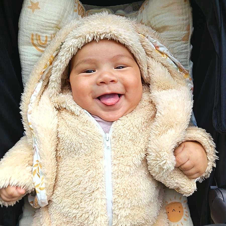 Kaelan a rejoint le concours — aidez-le/la à gagner de superbes lots ! apparel, baby, child, closeup, cozy, cushion, cute, face, fluffy_clothing, happy, hoodie, indoors, infant, patterned_textile, seated, smiling, tongue_out, warm_clothing, young_child, zipped_jacket