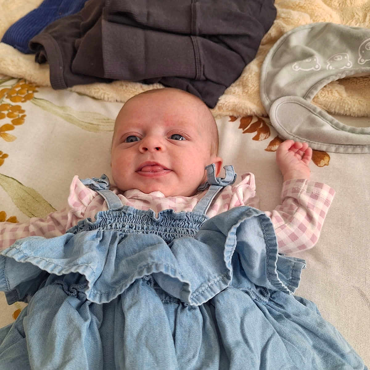 Tilly is registered to the contest to win money with this photo: baby, bed, blanket, clothing, dress, fabric, face, head, indoor, infant, infant_clothing, person, playful, portrait, ruffled_dress, shirt, soft_toy, textile, tongue_out, young_child