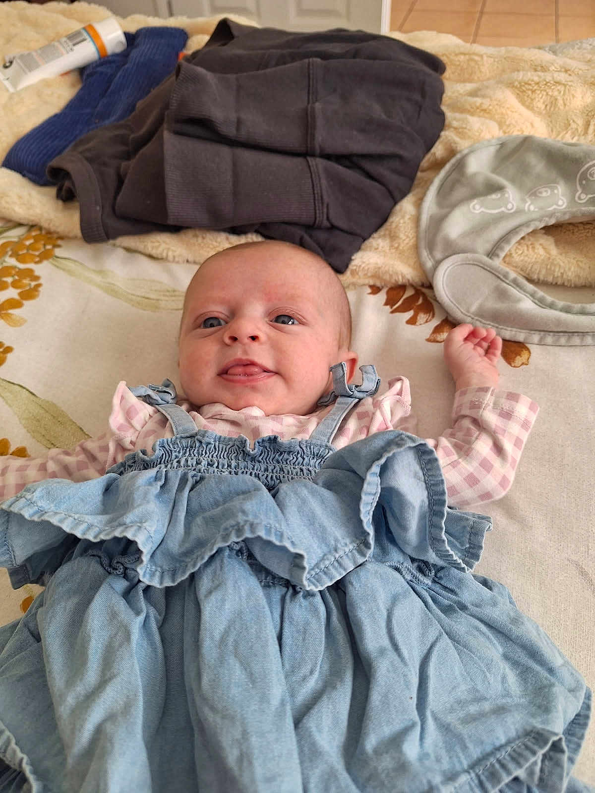 Tilly is registered to the contest to win money with this photo: baby, infant, bed, blanket, clothing, dress, fabric, face, head, indoor, infant_clothing, person, playful, portrait, ruffled_dress, shirt, soft_toy, textile, tongue_out, young_child