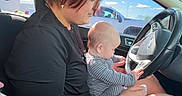 Elijah is registered to the contest to win money with this photo: adult, baby, bright, car, casual, daylight, driving, hand, inside, interaction, person, seat, short_hair, side_view, sitting, steering_wheel, striped_clothing, tattoo, vehicle, window