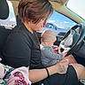 adult, baby, bright, car, casual, daylight, driving, hand, inside, interaction, person, seat, short_hair, side_view, sitting, steering_wheel, striped_clothing, tattoo, vehicle, window