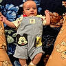 Elijah is registered to the contest to win money with this photo: baby, blanket, child, clothing, cozy, cute, expression, floral_pattern, footwear, indoors, infant, lying_down, mickey_mouse, pants, person, pluto, romper, sneakers, teddy_bear, wide_eyes