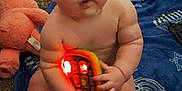 Declan is registered to the contest to win money with this photo: baby, child, toy, blanket, carpet, plush_toy, teddy_bear, playpen, indoor, floor, sitting, naked, face, hands, feet, light, red_light, blue_blanket, menorah_pattern, star_pattern