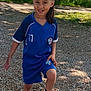 child, soccer_ball, blue_uniform, outdoor, gravel_path, greenery, sunlight, smiling, sport, footwear, shorts, jersey, nature, playing, happy, person, grass, daylight, casual, active
