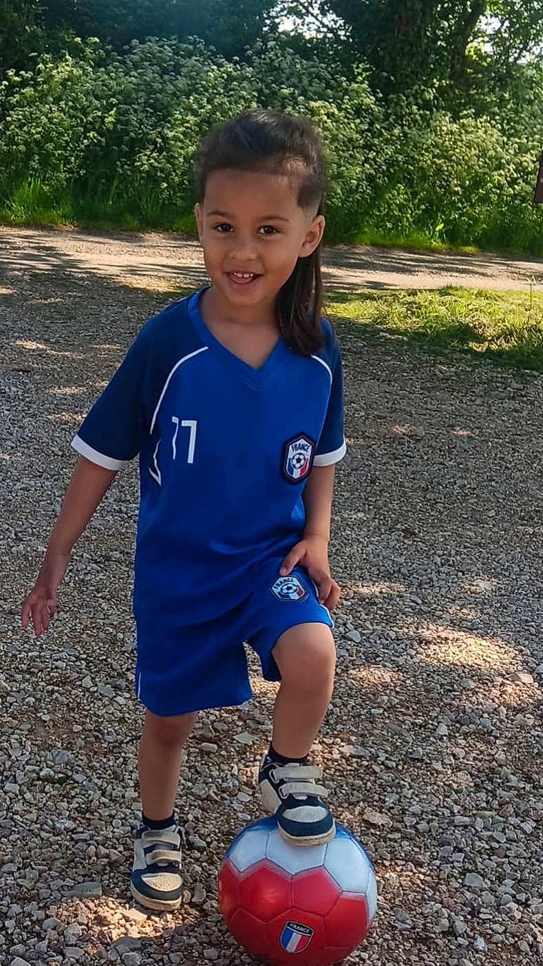 Tayden a rejoint le concours — aidez-le/la à gagner de superbes lots ! child, soccer_ball, blue_uniform, outdoor, gravel_path, greenery, sunlight, smiling, sport, footwear, shorts, jersey, nature, playing, happy, person, grass, daylight, casual, active
