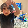 child, curly_hair, black_shirt, orange_shorts, living_room, carpet, toys, television, furniture, smile, happy, playful, person, indoor, kid, gesture, fun, home, young, curtain