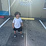 child, smiling, preschool, parking_lot, sunlight, lens_flare, water_bottle, white_tshirt, black_shorts, sneakers, outdoor, asphalt, yellow_parking_stop, door, window, building_wall, curly_hair, happy, standing, casual_clothing