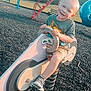 child, playground, spring_rocker, motorcycle_toy, smiling, blonde_hair, tshirt, shorts, sneakers, outdoor, park, trees, grass, slide, swing, bench, play_equipment, daylight, happy, fun