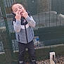 Tony a rejoint le concours — aidez-le/la à gagner de superbes lots ! child, boy, outdoor, fence, mesh_fence, rocks, casual_clothing, jacket, pants, shoes, playful, gesture, closed_eyes, standing, daylight, young, person, expression, fun, background