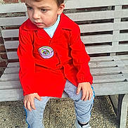 Tony participe au concours pour gagner de l'argent avec cette photo : child, boy, red_jacket, gray_pants, white_sneakers, bench, brick_wall, outdoor, pebbled_ground, casual_clothing, portrait, sitting, daylight, curious_expression, short_hair, young_child, footwear, person, street_style, relaxed_pose