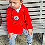 child, boy, red_jacket, gray_pants, white_sneakers, bench, brick_wall, outdoor, pebbled_ground, casual_clothing, portrait, sitting, daylight, curious_expression, short_hair, young_child, footwear, person, street_style, relaxed_pose