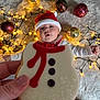 baby, santa_hat, cookie, hand, christmas_lights, ornaments, carpet, holiday, festive, red_clothing, white_clothing, decorations, indoors, cute, celebration, winter, smiling, child, face, holiday_cookie