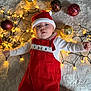 Cooper is registered to the contest to win money with this photo: baby, infant, christmas, holiday, santa_hat, red_overalls, string_lights, ornaments, gold_ornaments, red_ornaments, soft_texture, cute, festive, celebration, holiday_decor, face, person, lying_down, indoors, warm_lighting