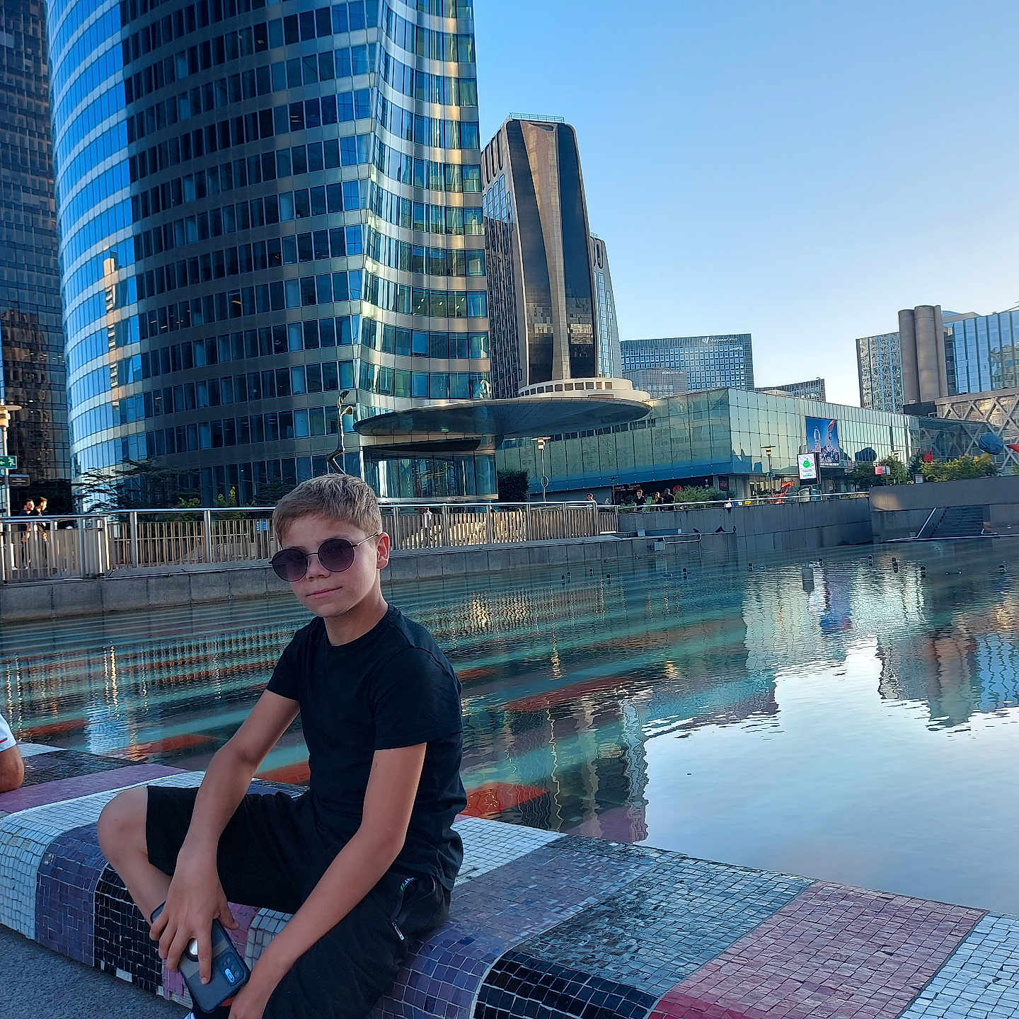 Ghenadie a rejoint le concours — aidez-le/la à gagner de superbes lots ! bench, black_clothing, boy, casual, child, city, daytime, glass_building, modern_architecture, mosaic, outdoor, phone, reflection, relaxing, shorts, skyscraper, sneakers, sunglasses, urban, water