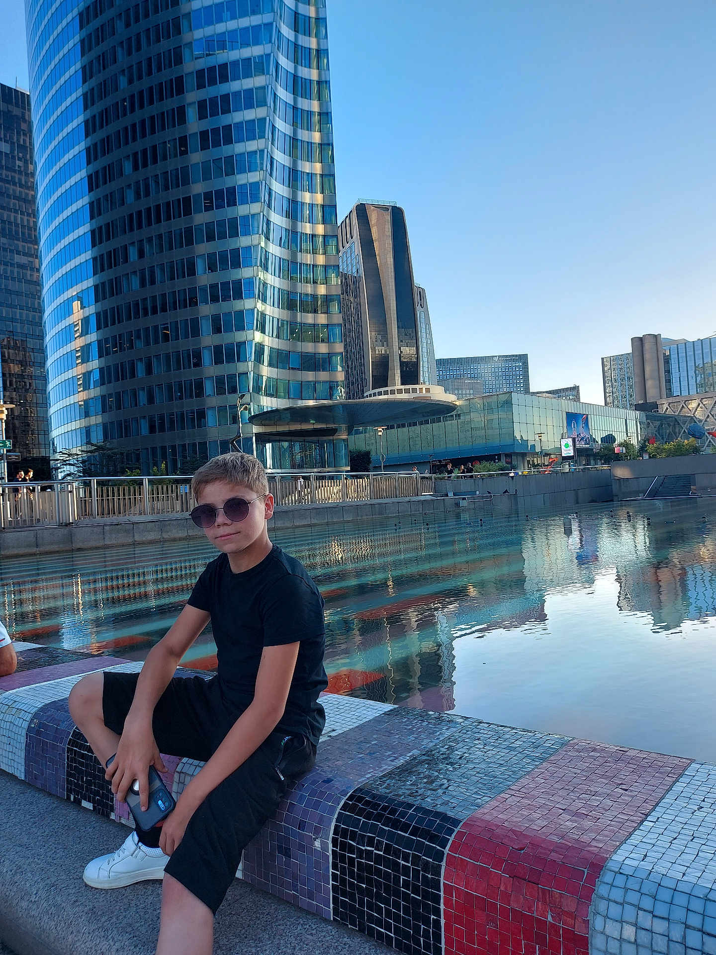 Ghenadie a rejoint le concours — aidez-le/la à gagner de superbes lots ! child, boy, sunglasses, black_clothing, shorts, sneakers, phone, bench, mosaic, water, reflection, urban, city, skyscraper, glass_building, daytime, outdoor, modern_architecture, casual, relaxing
