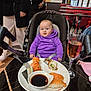 baby, child, sushi, plate, glass, drink, purple_hoodie, stroller, restaurant, food, table, people, indoor, fork, soy_sauce, seat, curious, casual_clothing, dining, asian_food