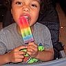 child, curly_hair, popsicle, rainbow, high_chair, smiling, happy, indoor, toddler, gray_shirt, food, snack, playful, heart_effects, cute, portrait, young_child, eating, colorful, fun