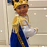 child, crown, costume, cape, blue, gold, smile, indoor, door, curly_hair, playful, portrait, young, person, floor, sign, white, fringe, gold_trim, happy