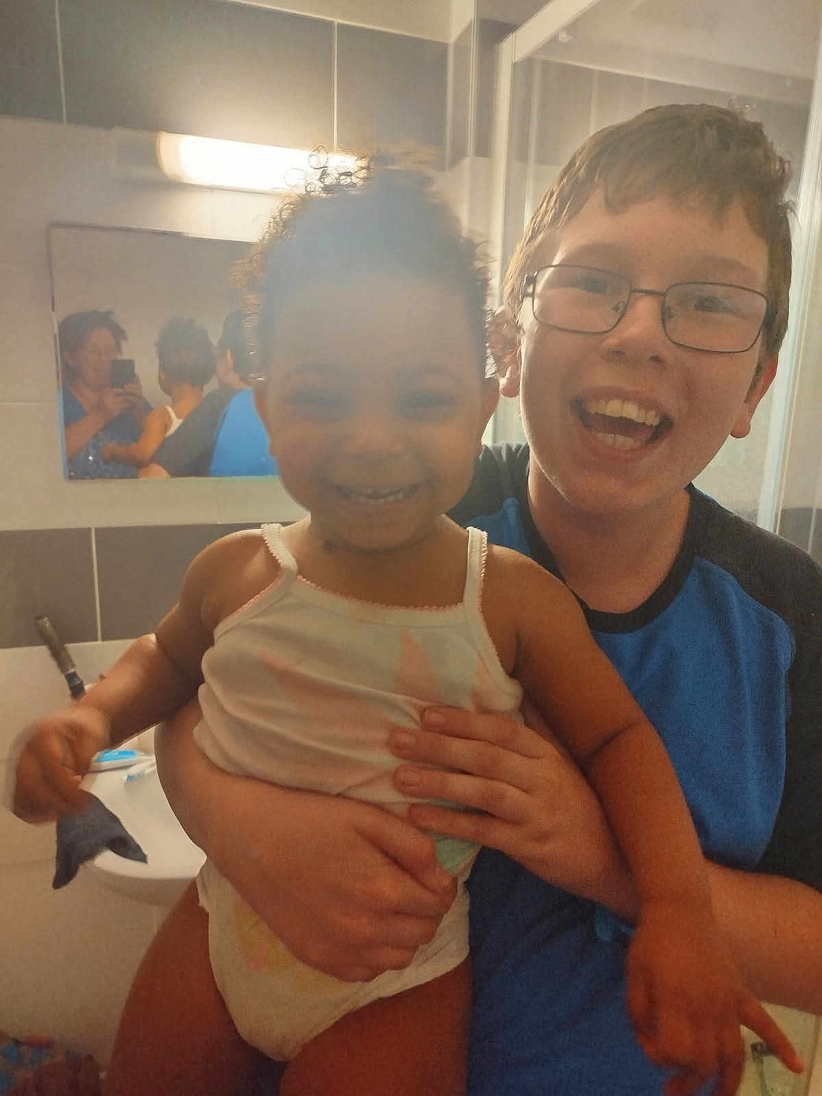 Noémie participe au concours pour gagner de l'argent avec cette photo : child, toddler, bathroom, mirror, glasses, smile, holding, person, sink, light, reflection, tank_top, diaper, happy, indoor, family, casual, curly_hair, short_hair, person_holding_child