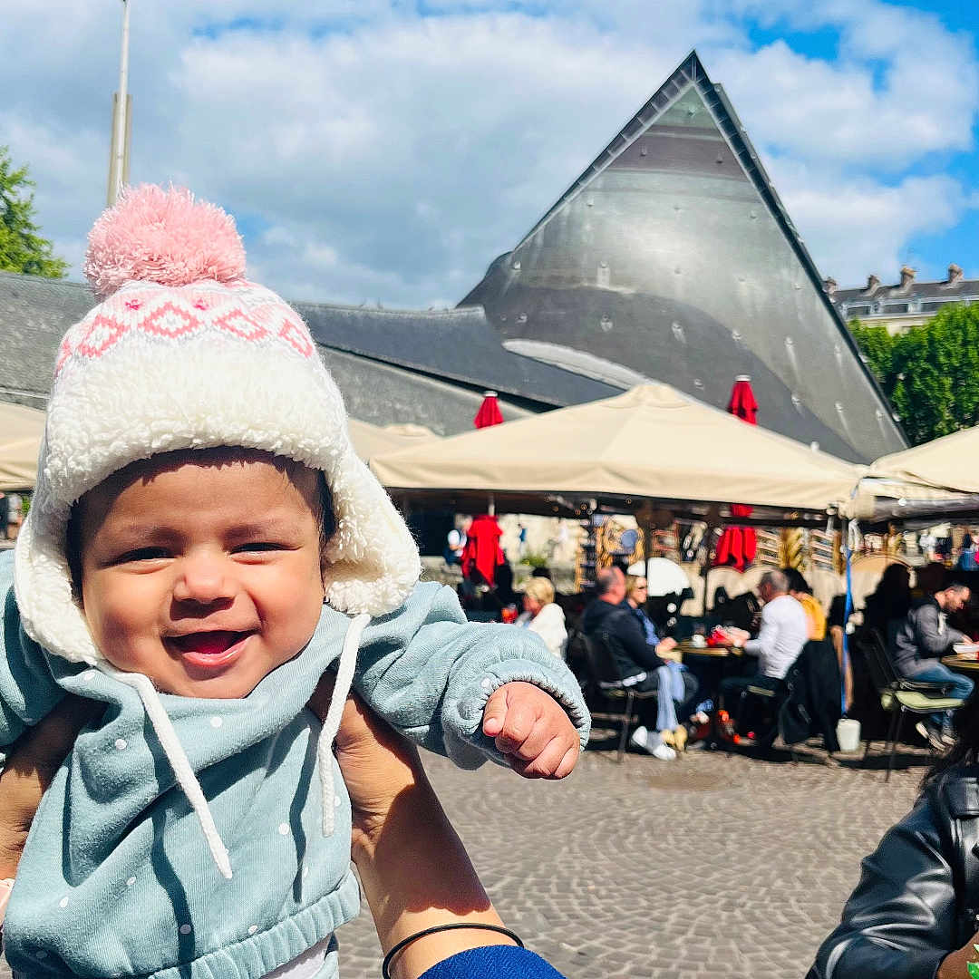 Kridha participe au concours pour gagner de l'argent avec cette photo : baby, blue_sky, cafe, child, clouds, daytime, hand, happy, hat, jacket, outdoor, pants, people, person, polka_dot, pom_pom, smiling, sunlight, table, wood