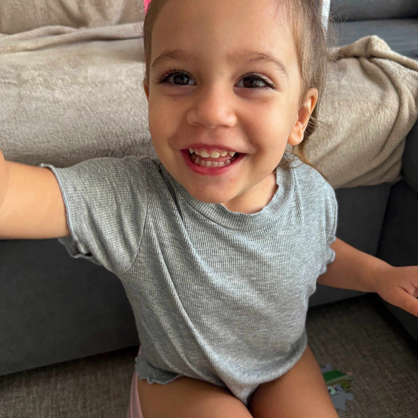 Djoulyna a rejoint le concours — aidez-le/la à gagner de superbes lots ! blanket, carpet, child, clothing, cute, face, grey_shirt, hair_tied, happy, home, indoors, joy, person, playful, shorts, sitting, smiling, sofa, toddler, young