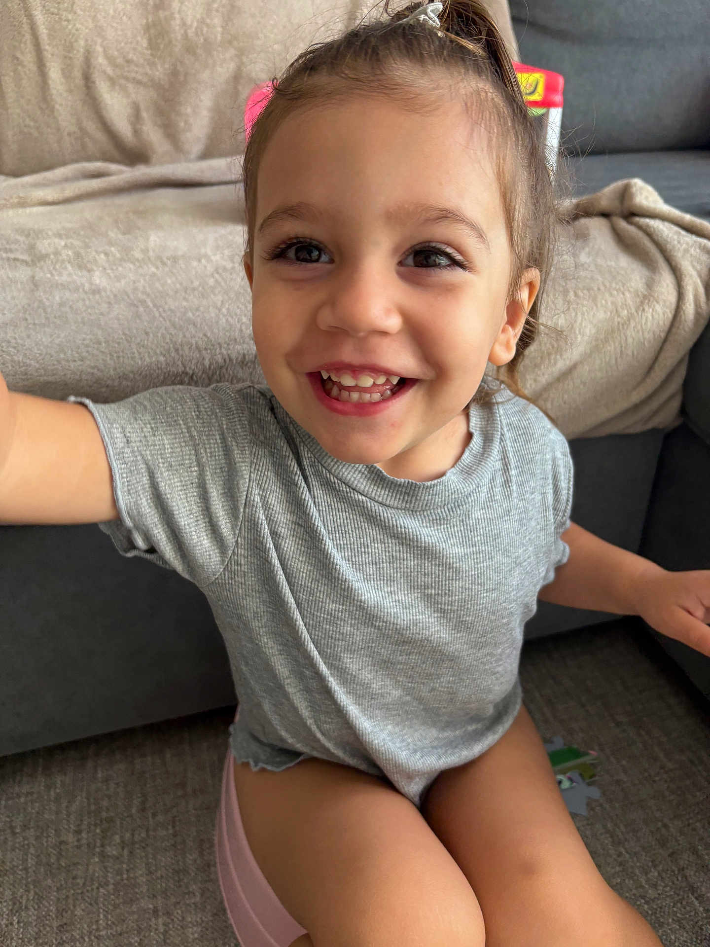 Djoulyna a rejoint le concours — aidez-le/la à gagner de superbes lots ! blanket, carpet, child, clothing, cute, face, grey_shirt, hair_tied, happy, home, indoors, joy, person, playful, shorts, sitting, smiling, sofa, toddler, young