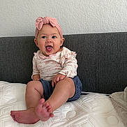 Mai joined the competition — help win amazing prizes! baby, child, infant, headband, pink_bow, smiling, happy, sitting, mattress, foot, clothing, long_sleeve, shorts, indoor, wall, couch, texture, person, cute, playful