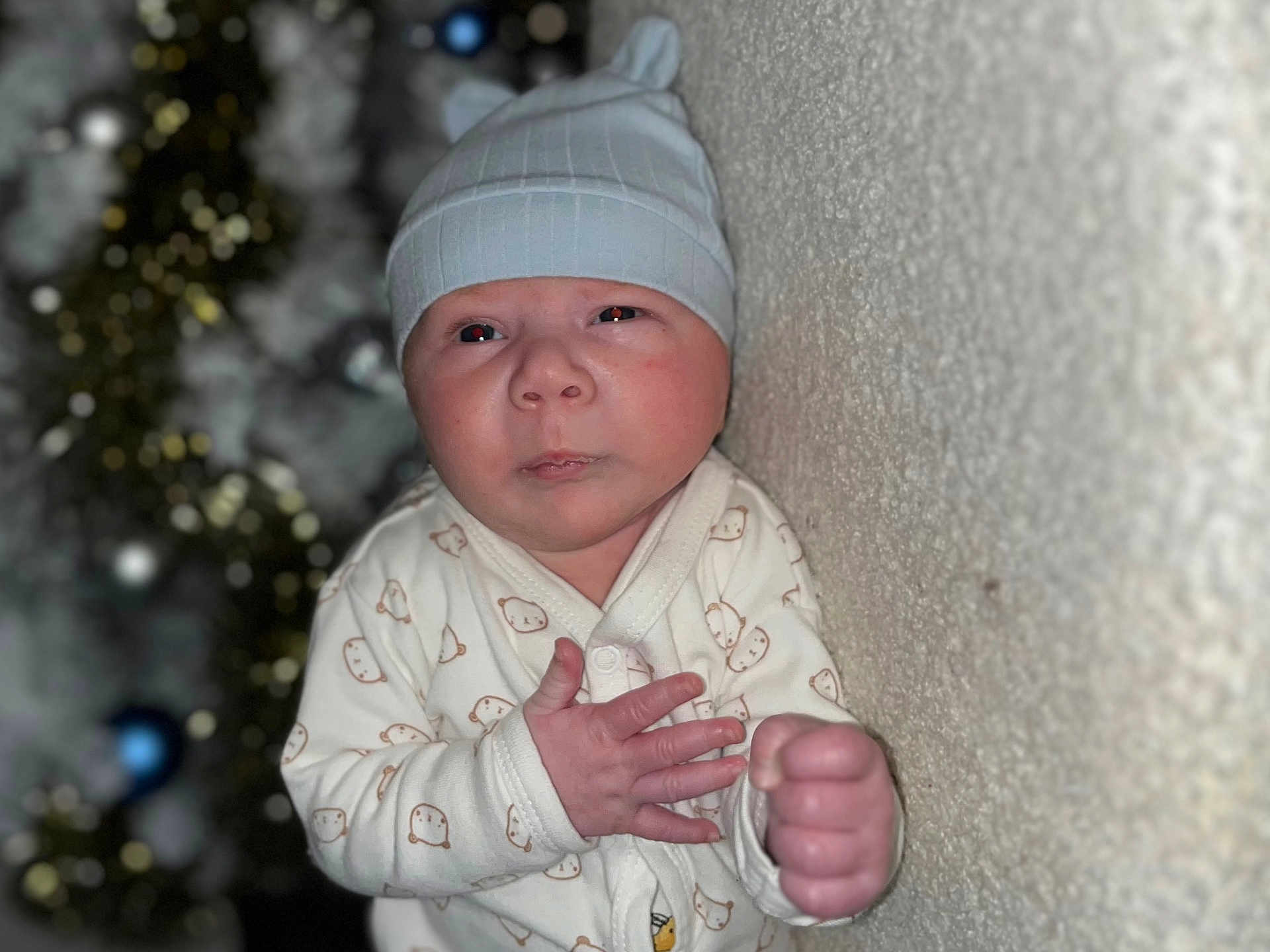 Tylio a rejoint le concours — aidez-le/la à gagner de superbes lots ! baby, bear_pattern, blurred_background, child, cute, decorations, expression, face, fist, hand, hat, holiday, indoor, infant, lights, newborn, onesie, portrait, texture, wall