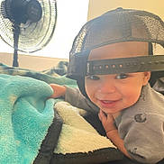 Jayden joined the competition — help win amazing prizes! child, toddler, baby, smiling, portrait, indoor, bedroom, blanket, cozy, baseball_cap, backwards_hat, fan, bedding, gray_shirt, close_up, happy, face, sitting, home, soft_lighting