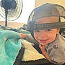 child, toddler, baby, smiling, portrait, indoor, bedroom, blanket, cozy, baseball_cap, backwards_hat, fan, bedding, gray_shirt, close_up, happy, face, sitting, home, soft_lighting