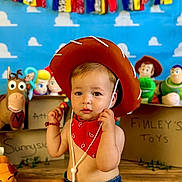 Finley is registered to the contest to win money with this photo: baby, child, cowboy_hat, red_bandana, toy_story, toys, indoor, floor, wooden_floor, colorful_background, blue_background, stuffed_toys, smiling, portrait, playful, young_child, cute, decorations, sitting, costume