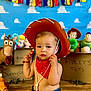baby, child, cowboy_hat, red_bandana, toy_story, toys, indoor, floor, wooden_floor, colorful_background, blue_background, stuffed_toys, smiling, portrait, playful, young_child, cute, decorations, sitting, costume