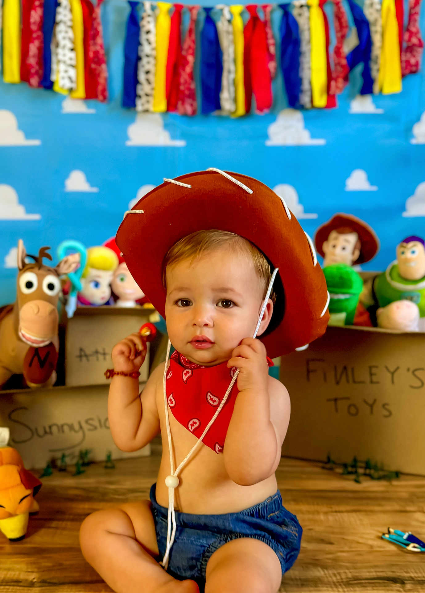 Finley is registered to the contest to win money with this photo: baby, child, cowboy_hat, red_bandana, toy_story, toys, indoor, floor, wooden_floor, colorful_background, blue_background, stuffed_toys, smiling, portrait, playful, young_child, cute, decorations, sitting, costume