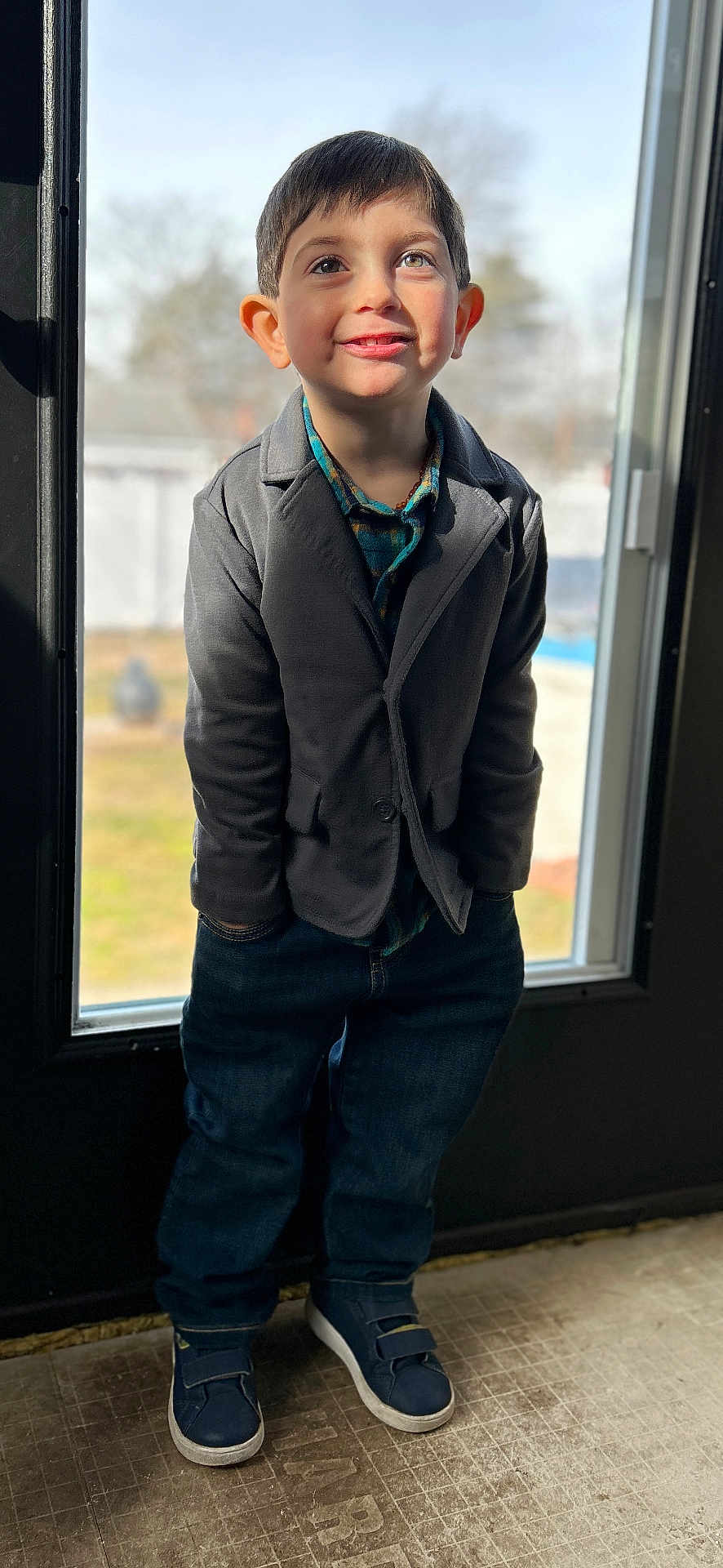 Elliot is registered to the contest to win money with this photo: child, boy, smiling, standing, indoor, window, door, casual_clothing, jeans, sneakers, blazer, plaid_shirt, hands_in_pockets, short_hair, daylight, portrait, happy, young, casual, bright
