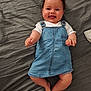 baby, smiling, denim_dress, white_shirt, lying_down, gray_sheet, happy, infant, cute, portrait, child, skin, clothing, small_hands, feet, indoors, one_person, young_child, joyful, casual