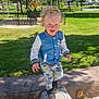 child, curly_hair, daylight, fence, grass, happy, jacket, leaves, log, mickey_mouse, nature, outdoor, pants, park, playground, shoes, smiling, sunny, toddler, trees