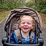 baby_stroller, blue_shirt, child, curly_hair, daylight, forest, greenery, happy, leaves, nature, outdoor, pants, path, person, seatbelt, smiling, stroller, toddler, trees, walking_path