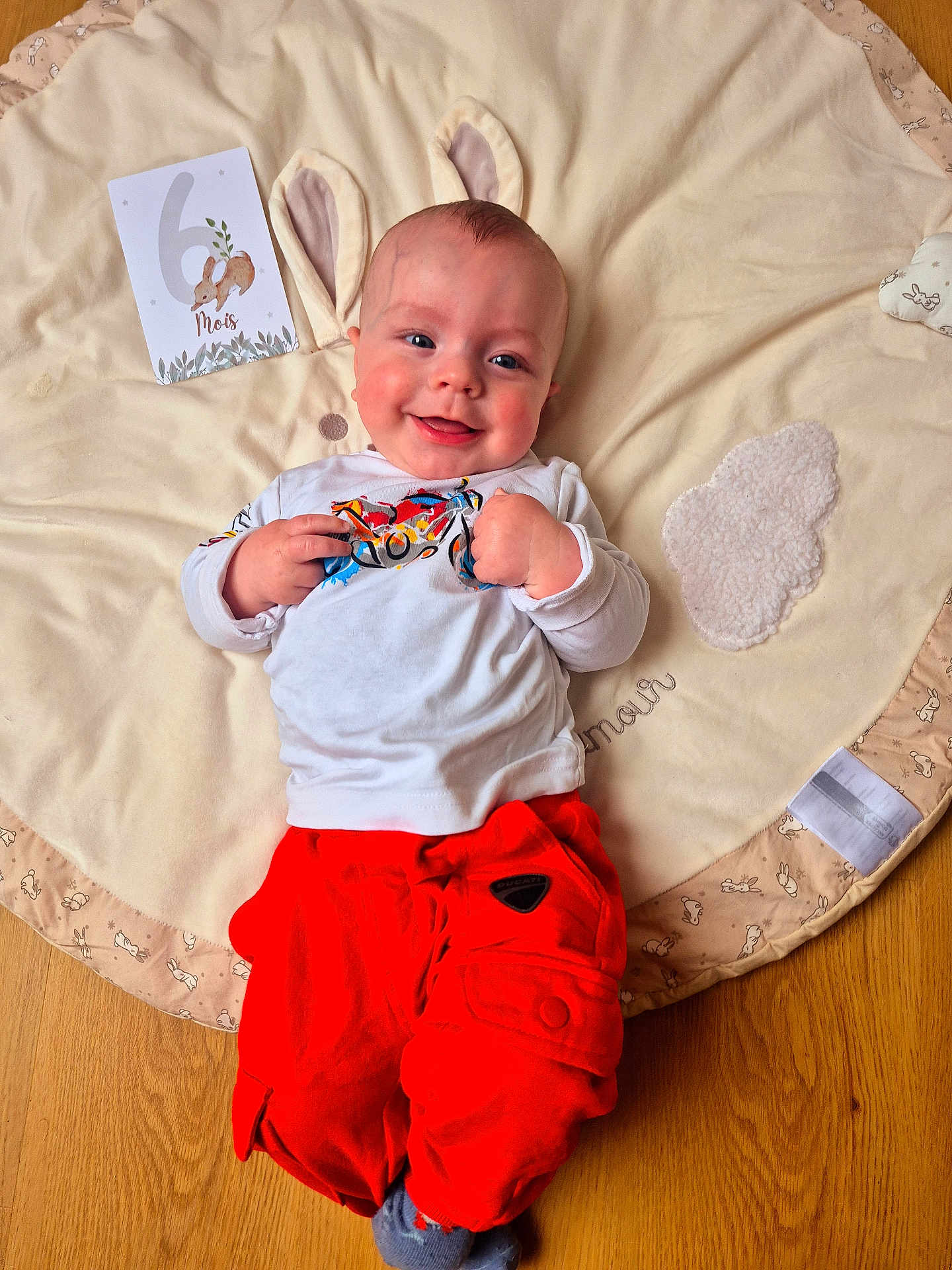 Abel a rejoint le concours — aidez-le/la à gagner de superbes lots ! baby, infant, smiling, happy, playmat, blanket, milestone_card, bunny_ears, red_pants, white_shirt, socks, hardwood_floor, portrait, rosy_cheeks, card, lying_down, cozy, cute, clothing, face
