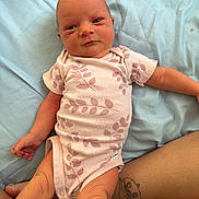 Elaine joined the competition — help win amazing prizes! baby, newborn, infant, onesie, pink_clothing, leaf_pattern, tattoo, human_leg, light_blue_sheet, lying_down, skin, person, cute, small_hands, small_feet, infant_face, resting, indoors, soft_light, casual