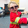 Ryder joined the competition — help win amazing prizes! child, sunglasses, red_jacket, reflective_stripes, table, office_chair, door, carpet, indoor, playful, smiling, person, short_hair, young_child, furniture, mischievous, curly_hair, black_chair, table_edge, casual