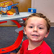 Ryder is registered to the contest to win money with this photo: child, boy, smiling, red_shirt, hot_wheels, toy, race_track, indoor, carpet, door, play, happy, thumbs_up, face, person, technology, phone_screen, app_icons, weather_alert, background