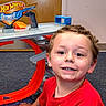child, boy, smiling, red_shirt, hot_wheels, toy, race_track, indoor, carpet, door, play, happy, thumbs_up, face, person, technology, phone_screen, app_icons, weather_alert, background