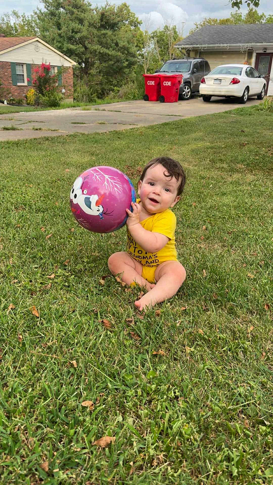 Christian joined the competition — help win amazing prizes! baby, child, grass, ball, outdoor, toy, playing, smiling, curly_hair, yellow_onesie, driveway, car, house, suburban, daytime, nature, greenery, happy, sitting, person